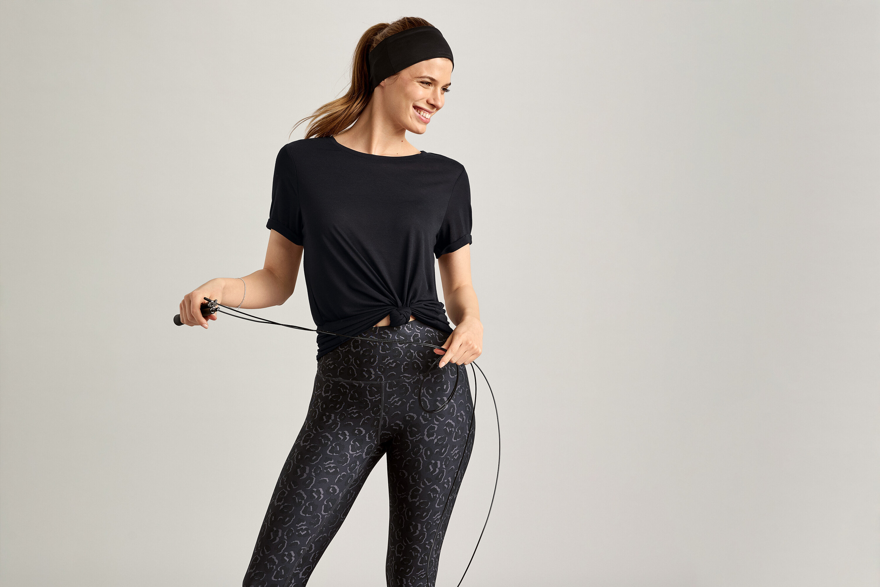 Frau mit Stirnband und Sportshirt, schwarz, hält ein Springseil vor grauem Hintergrund.