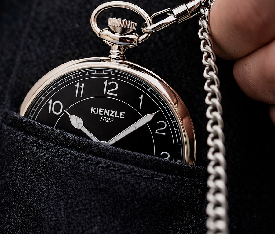 Nahaufnahme einer Person, die eine silberne Taschenuhr mit schwarzem Zifferblatt und Kette in einer dunklen Stofftasche hält.