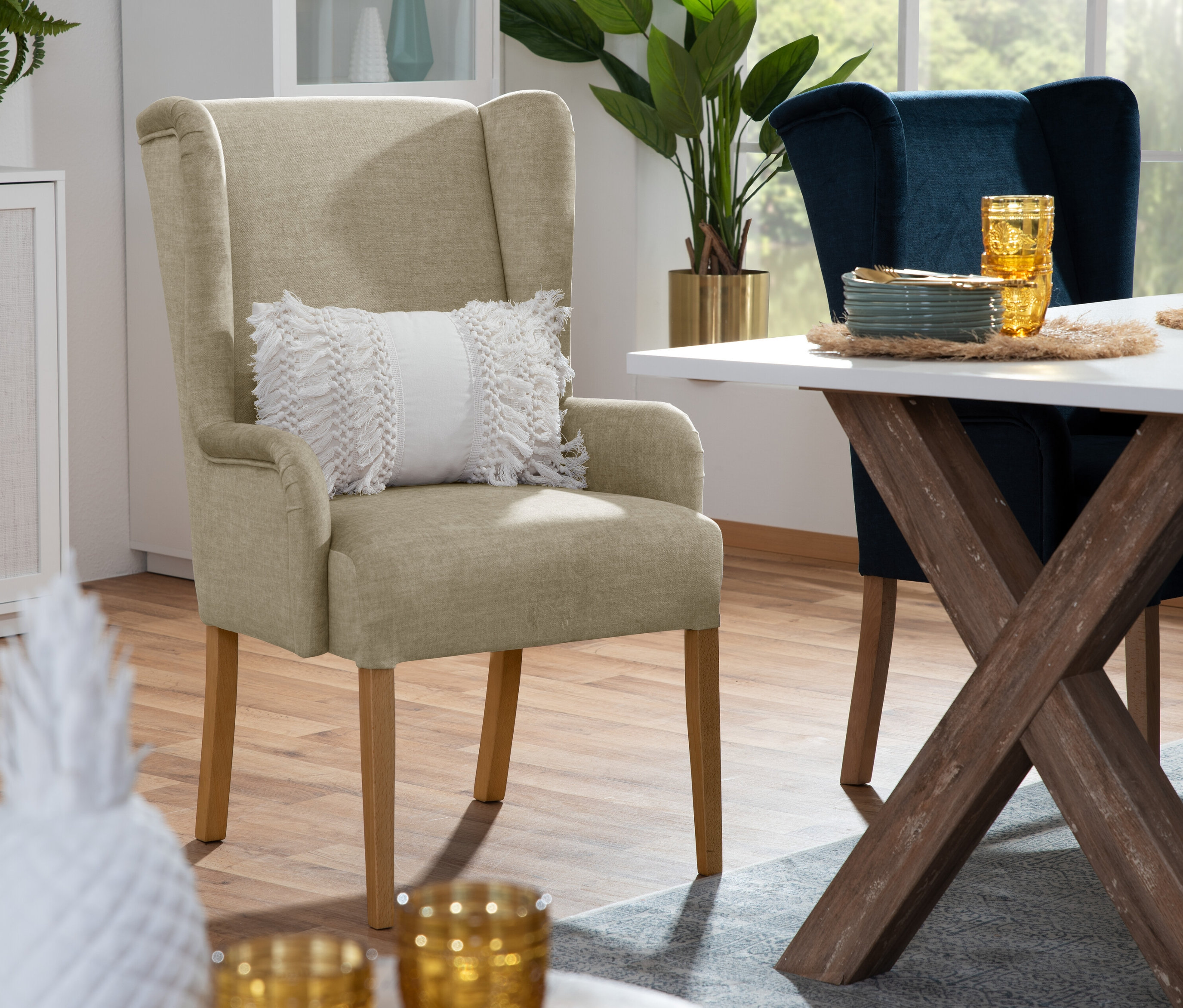 Beiger Hochlehner-Polsterstuhl »Hendrick« mit Armlehne und Kissen steht neben Esstisch mit Geschirr und blauem Stuhl.