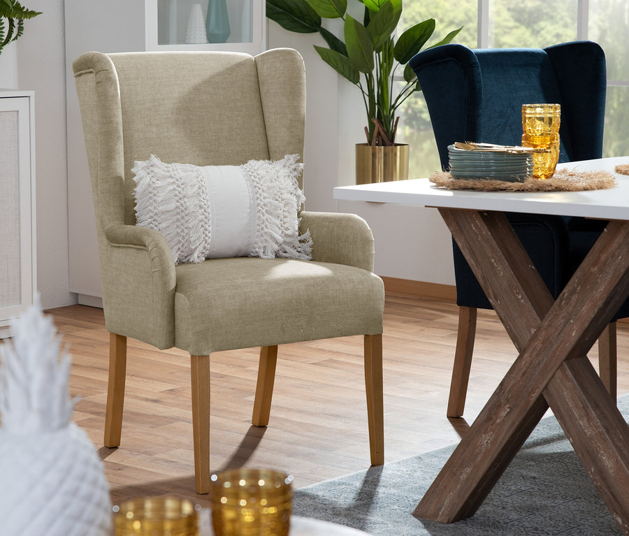 Beiger Hochlehner-Polsterstuhl »Hendrick« mit Armlehne und Kissen steht neben Esstisch mit Geschirr und blauem Stuhl.