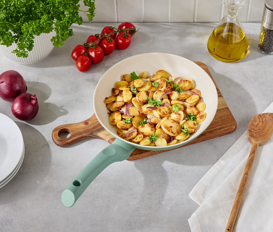Bratkartoffeln mit Zwiebeln und Petersilie in einer hellen Pfanne auf einem Holztisch mit Beilagen.