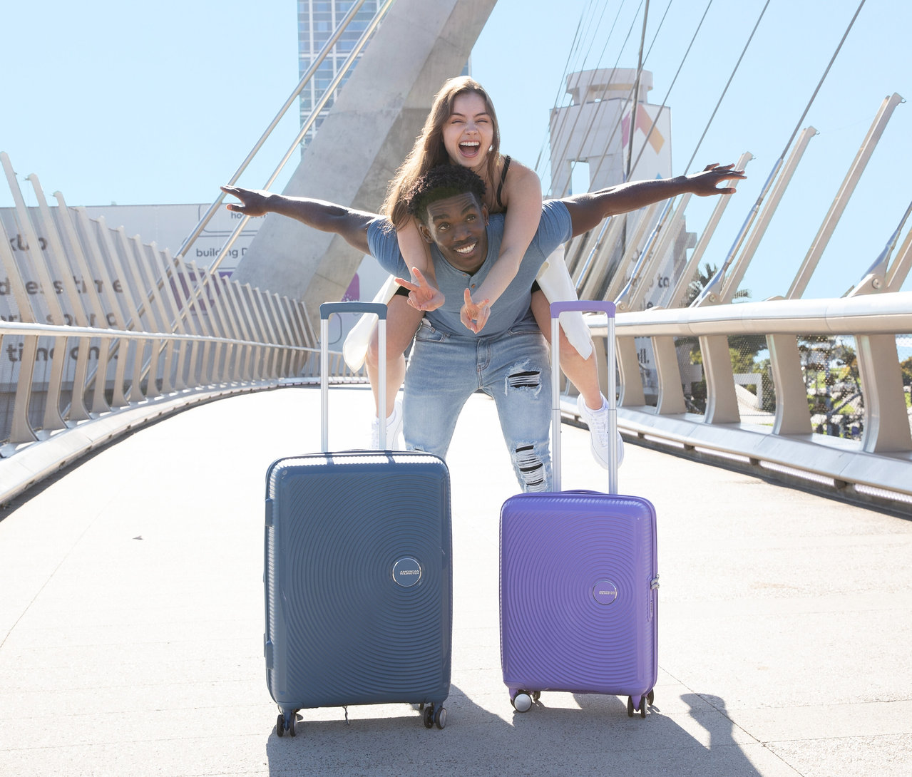 Mann mit Frau huckepack neben American Tourister Hartschalen-Koffer »Soundbox« Spinner, klein, lavender und groß, stone blue.