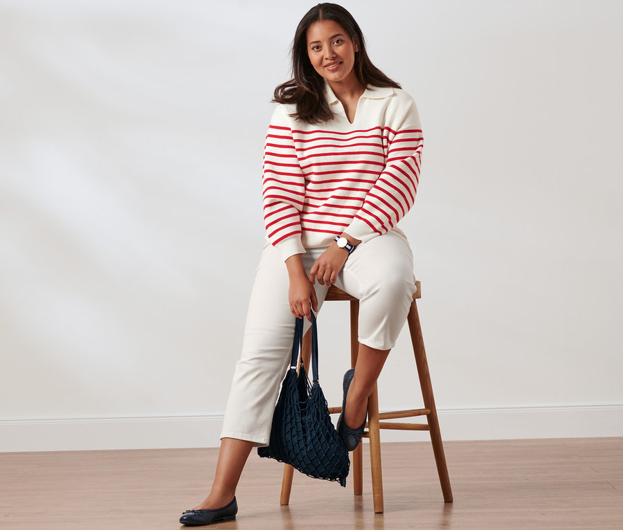 Eine Frau sitzt auf einem Holzhocker und trägt einen weiß-roten Strickpullover mit Kragen, Slimfit-Jeans, Leder-Ballerinas und eine Quarz-Armbanduhr aus Edelstahl. Sie hält eine Netztasche.