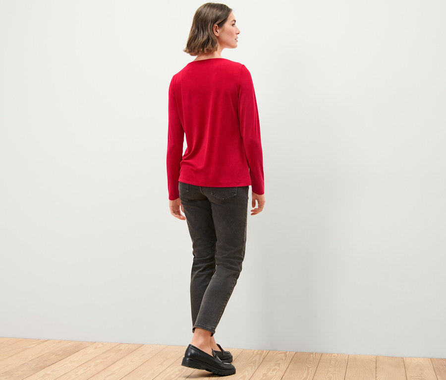 Frau steht mit dem Rücken zur Kamera und trägt ein Blusenshirt mit Raffung, Jeans und Schuhe.