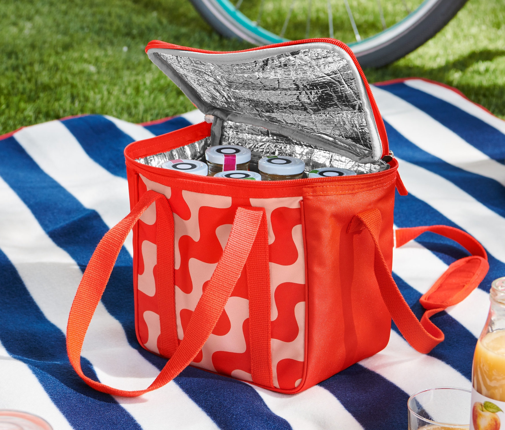 Eine offene kleine Kühltasche mit rot-weissem Muster steht auf einer blau-weiss gestreiften Decke, gefüllt mit Gläsern. Im Hintergrund sind ein Fahrrad und ein Helm zu sehen.