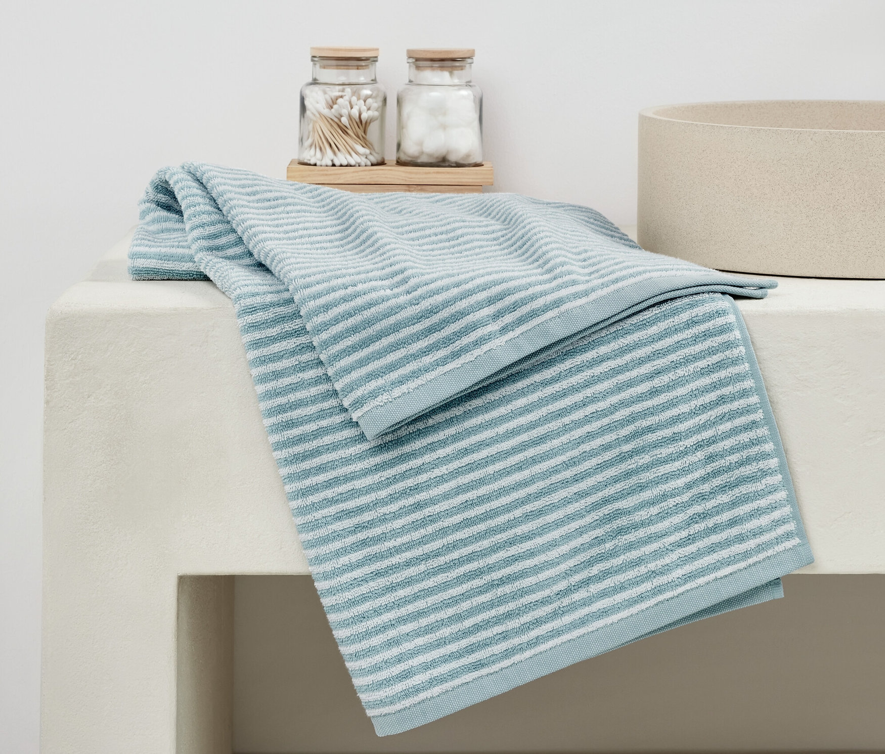 Ein hochwertiges blaues Badetuch liegt gefaltet auf einem weißen Waschbecken neben Gläsern mit Wattestäbchen und -bällchen.