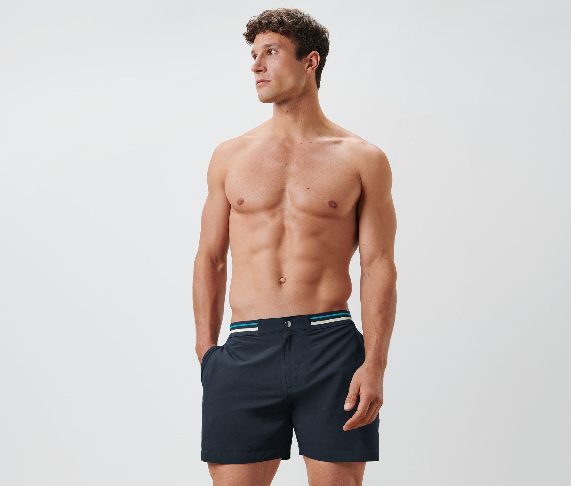 Mann mit freiem Oberkörper posiert in dunkelblauen Badeshorts.
