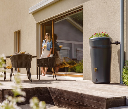 Frau steht mit Getränk auf Terrasse. Im Hintergrund ist ein 2-in-1-Regentonne-Komplett-Set »Azura«, ca. 230 l.