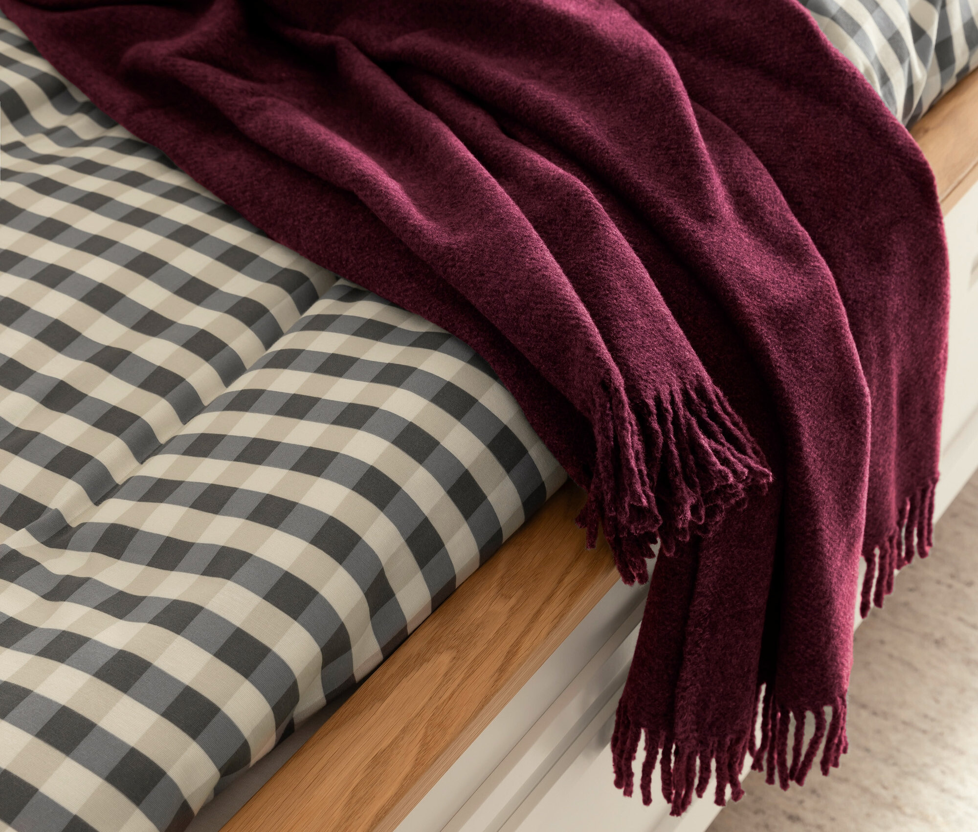Eine weinrote Decke mit Fransen liegt auf einem Bett mit Karomuster.