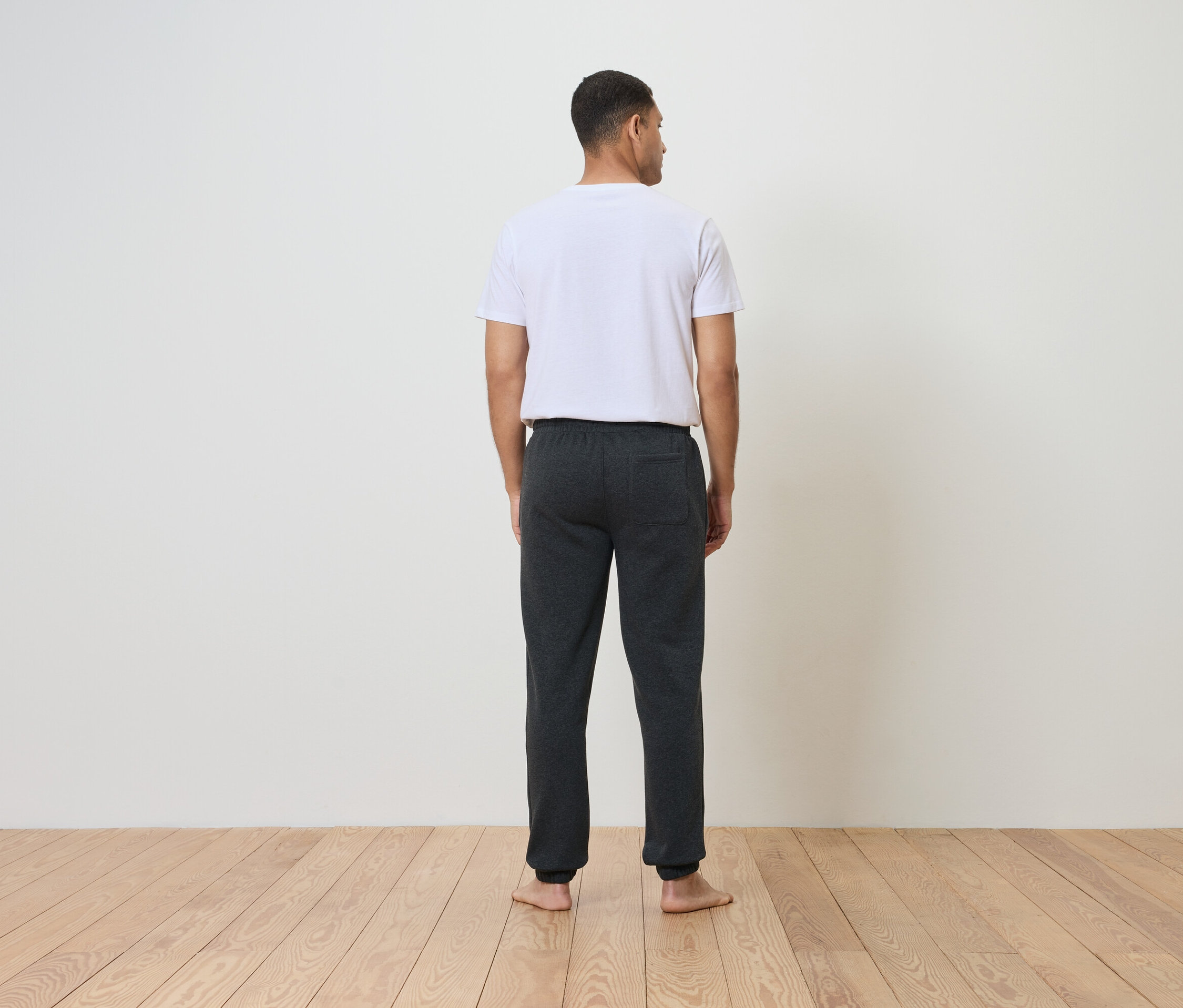 Mann mit weißem T-Shirt und dunkler Trainingshose steht mit dem Rücken zur Kamera auf einem Holzboden.