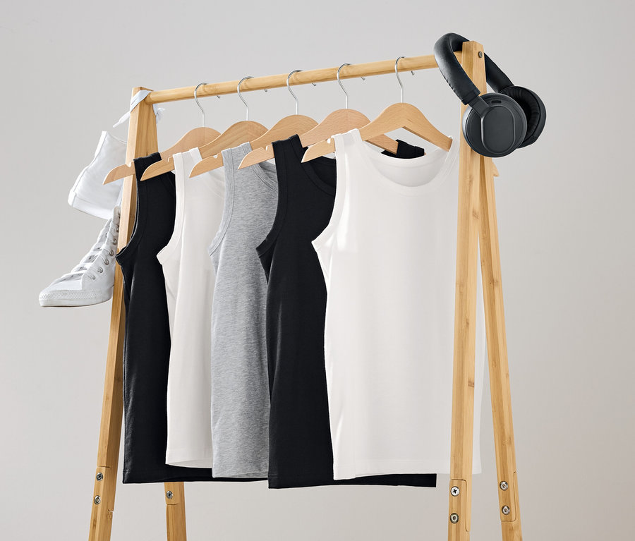 An einer hölzernen Kleiderstange hängen Tanktops in Schwarz, Weiß und Grau. Ein weißer Schuh und schwarze Kopfhörer hängen an der Stange.
