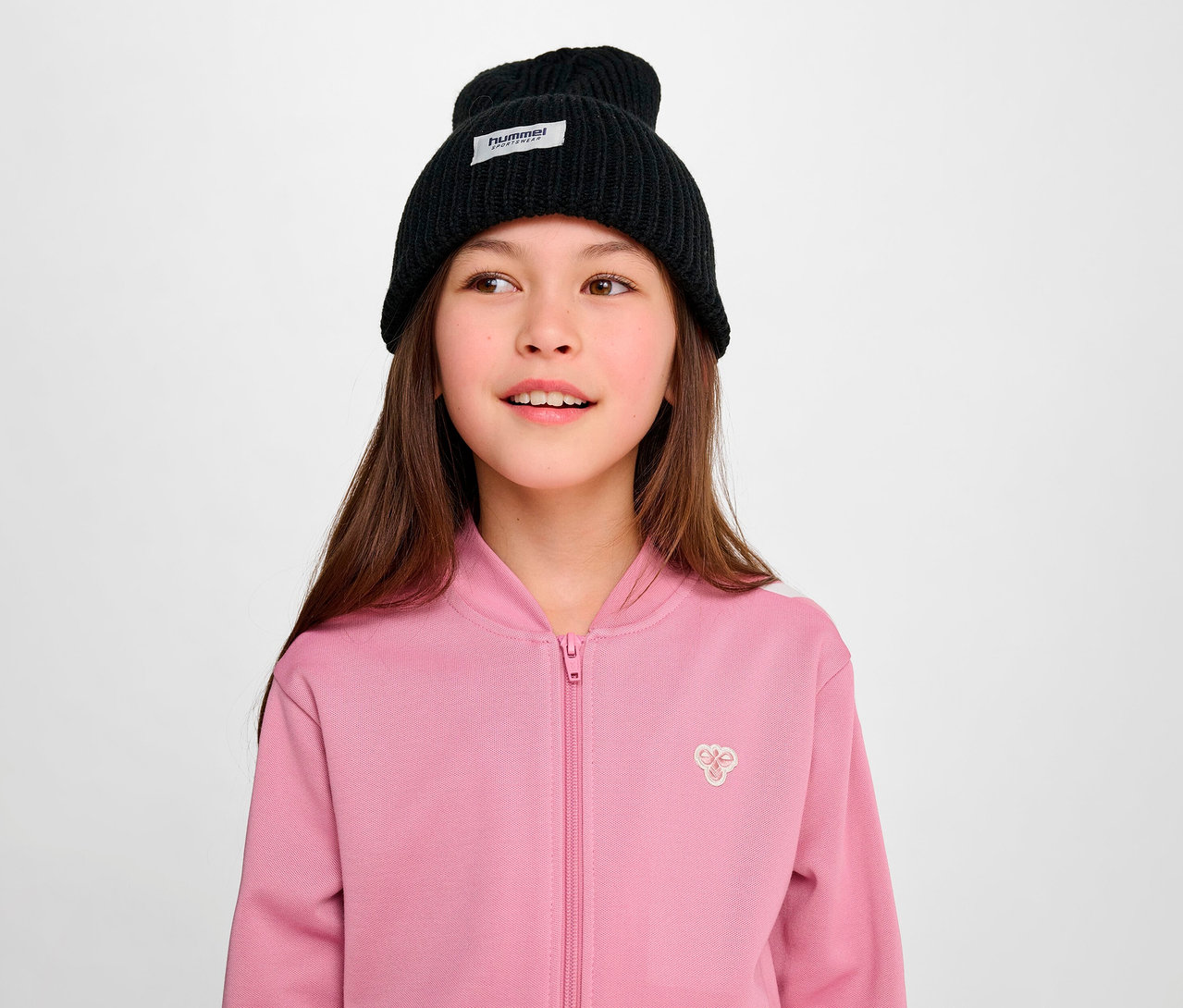 Junges Mädchen trägt eine schwarze Mütze und eine rosa Hummel HMLJR Archive Reg Poly Zip Jacket.