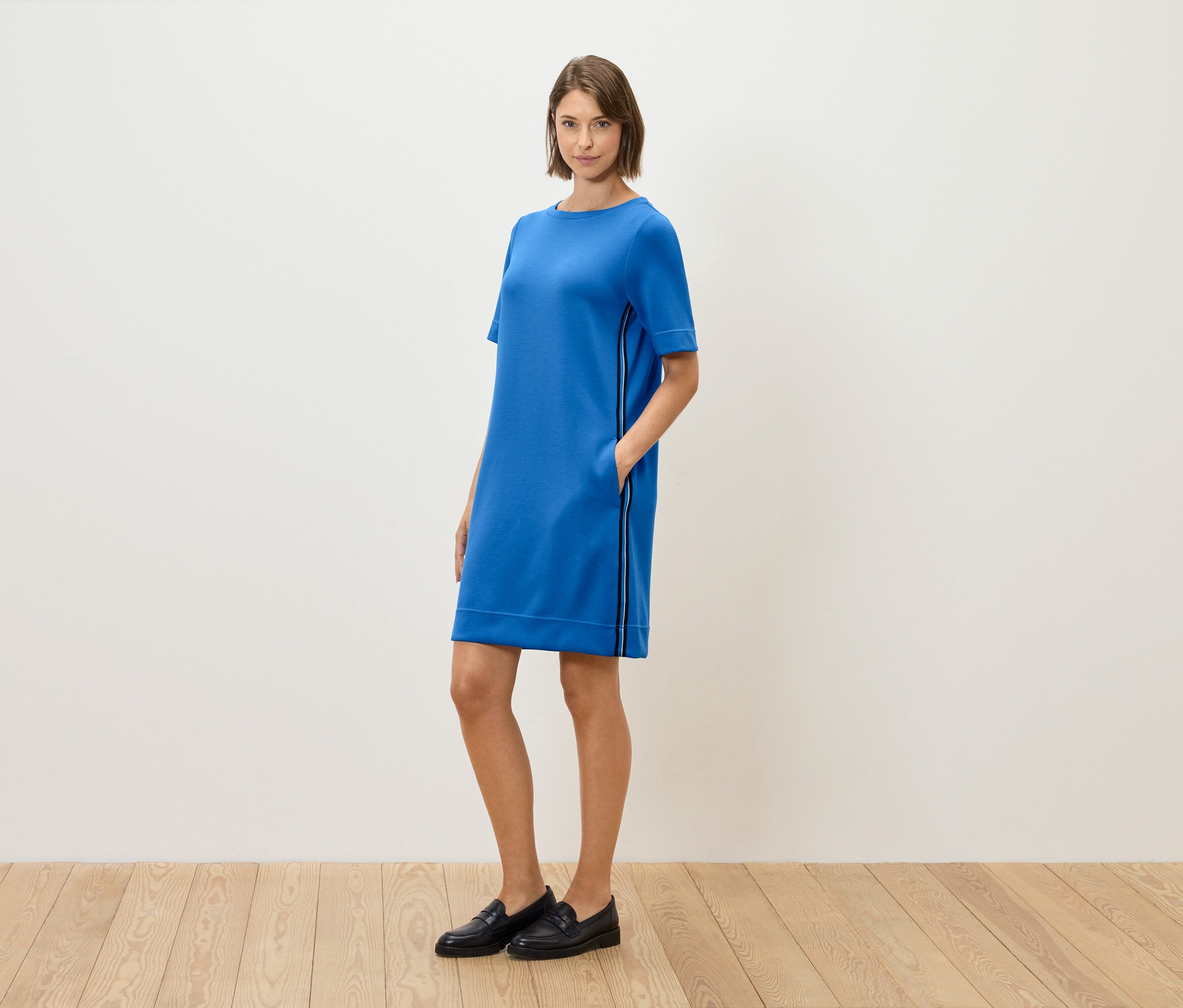 Eine Frau steht und posiert in einem blauen, kurzärmeligen Kleid und schwarzen Loafers.
