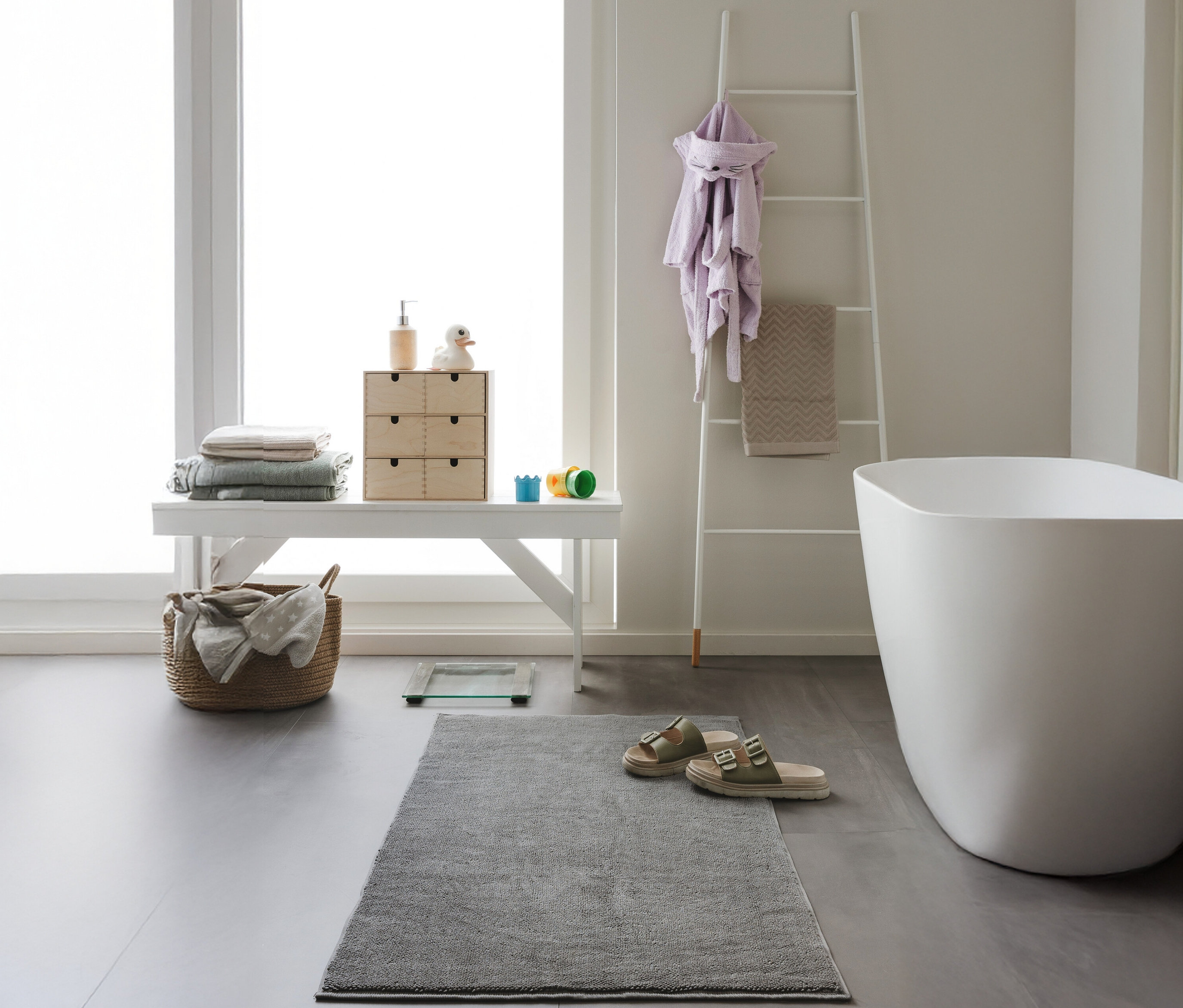 Ein anthrazitfarbener, rechteckiger Badvorleger »Ole« von benuta (70 x 120 cm) liegt vor einem weißen Hocker mit Handtüchern in einem Badezimmer.