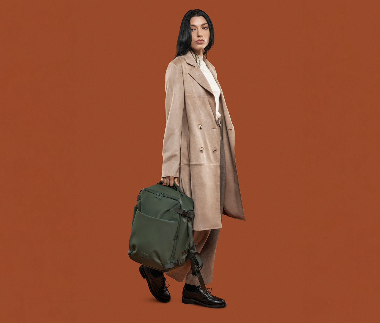 Frau mit beigem Mantel hält einen grünen Tucano TUGO DRY Kabinenrucksack mit 15,6"-Laptopfach.