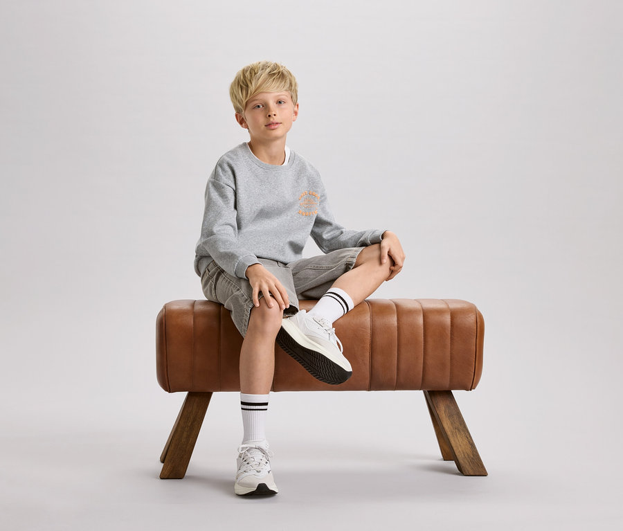 Ein blonder Junge sitzt auf einem braunen Turnbock. Er trägt ein graues Sweatshirt, Shorts und weiße Socken mit schwarzen Streifen.