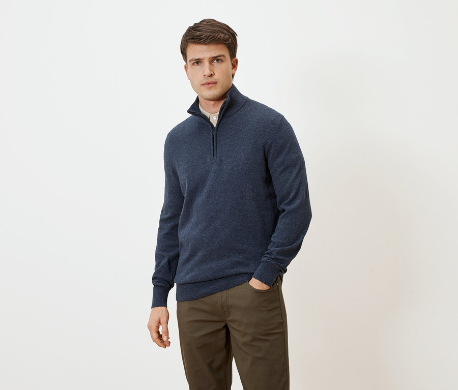 Mann mit blauem Pullover mit Reißverschluss und brauner Hose, eine Hand in der Hosentasche, vor einem neutralen Hintergrund.