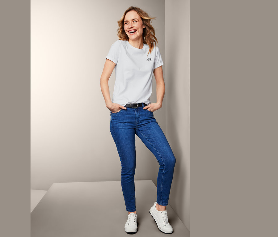 Eine lachende Frau steht in einem weißen Shirt mit kleiner Statement-Stickerei, blauen Jeans und weißen Schuhen.