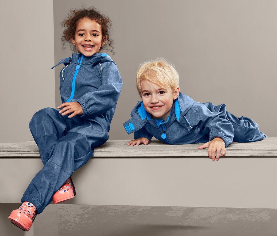 Zwei Kinder, ein Mädchen sitzt und ein Junge liegt auf einer Bank in blauen Softshelloveralls.