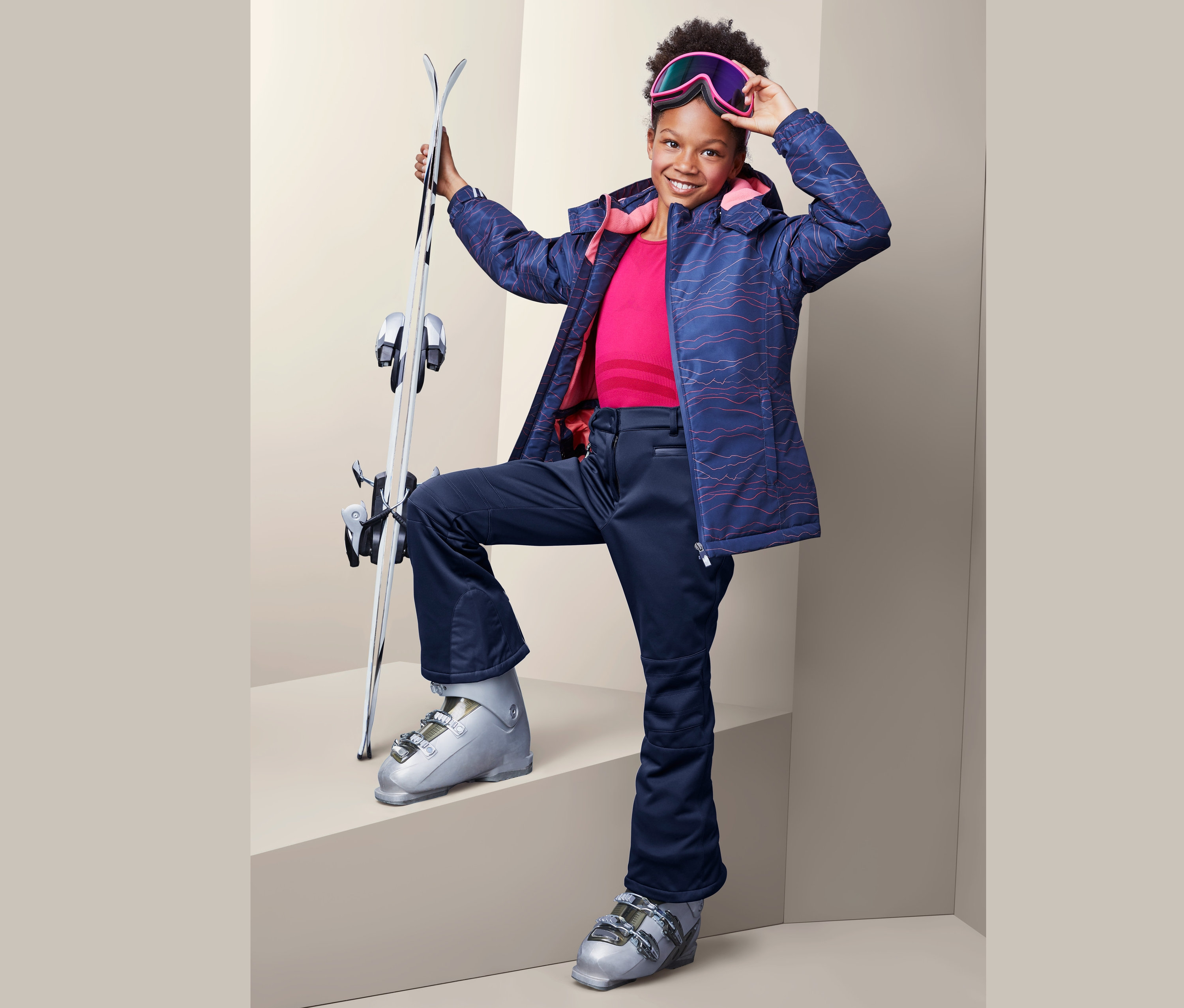 Lächelndes junges Mädchen posiert mit Skiern, trägt eine Jetskihose, eine Jacke und eine Skibrille.