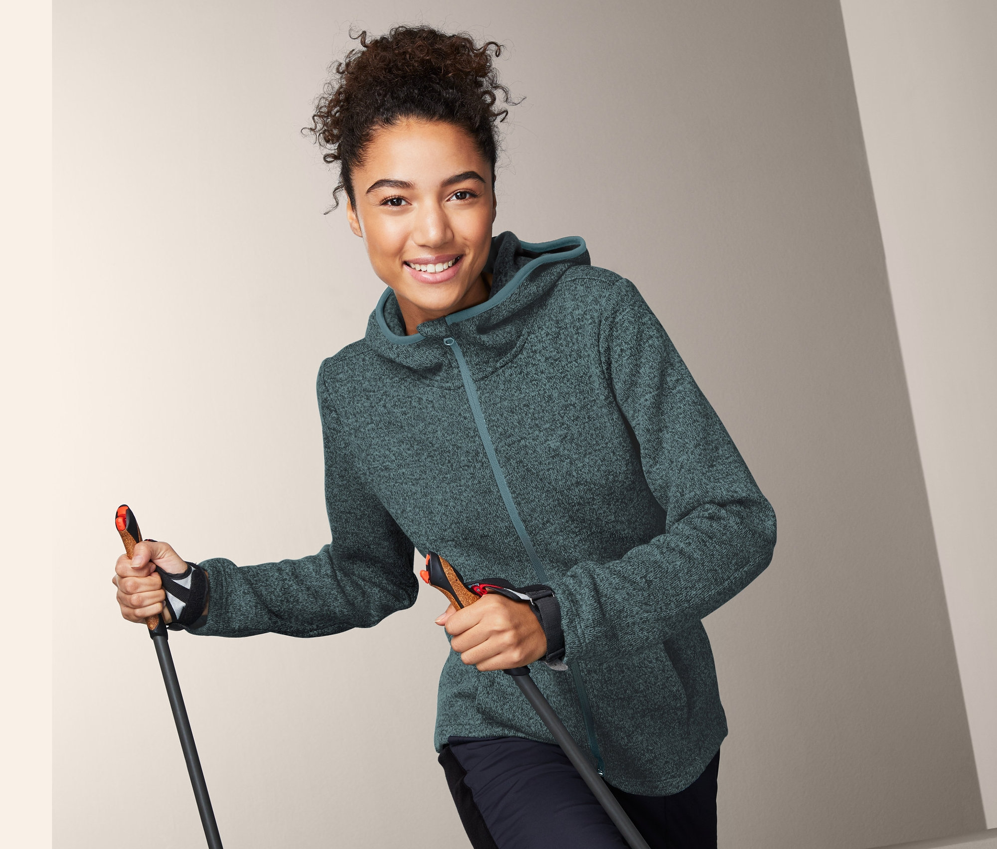 Lächelnde Frau hält Skistöcke und trägt eine Strickfleecejacke mit Kapuze.