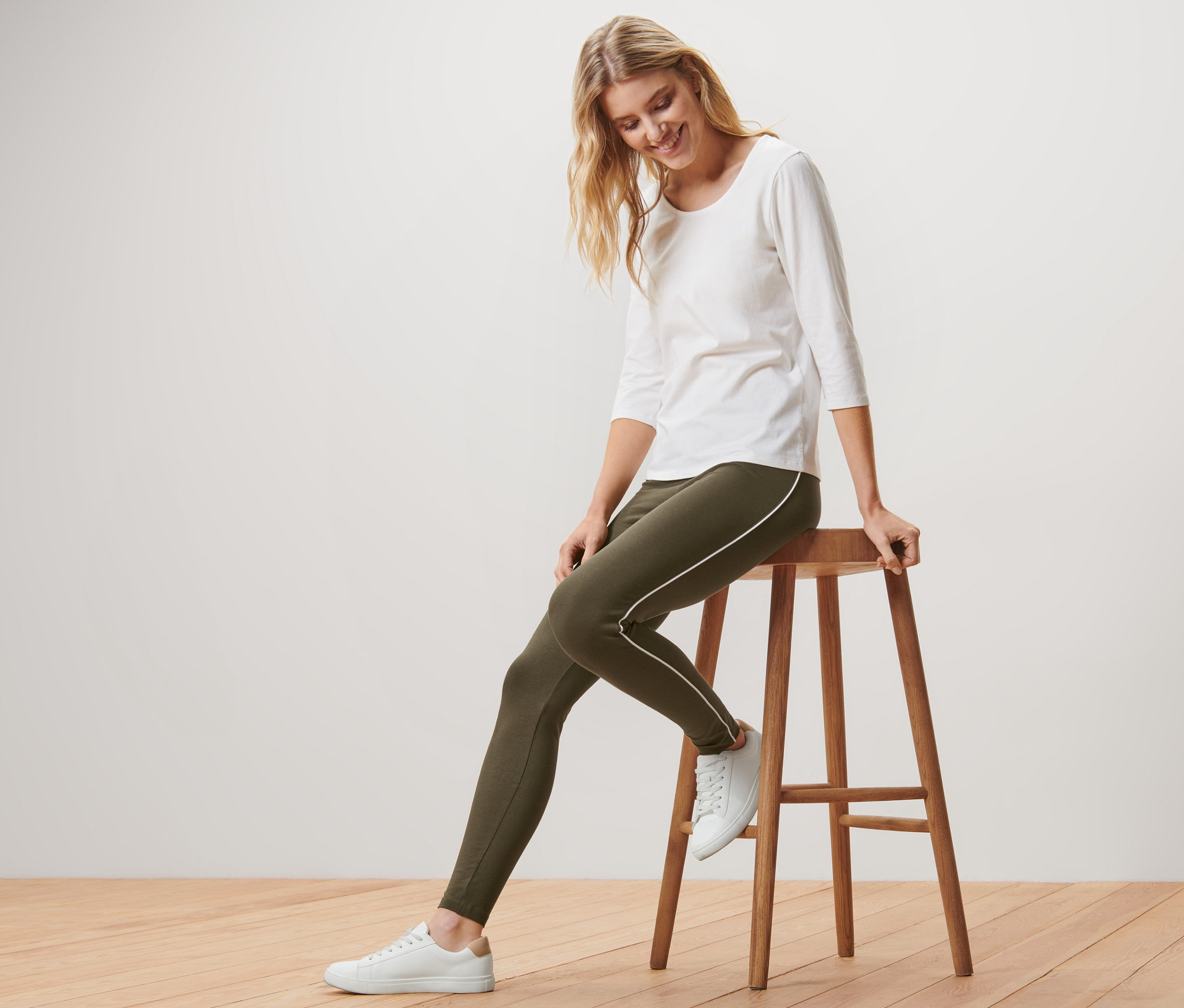 Blonde Frau sitzt auf einem Holzhocker und trägt grüne Leggings mit Paspelierung, ein weißes Shirt und Turnschuhe.