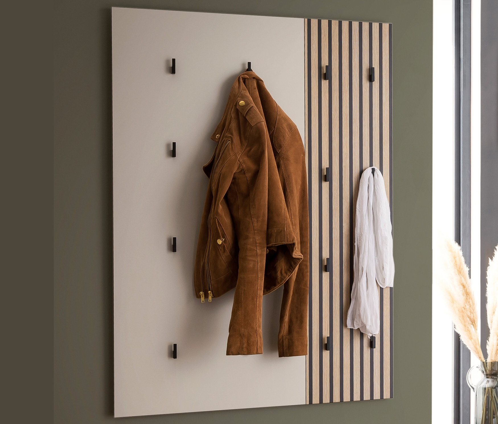 Eine braune Jacke hängt an einem Hakenpaneel »Alexa«, daneben ein Schal. Auf einer Bank liegen ein Kissen und eine Decke.