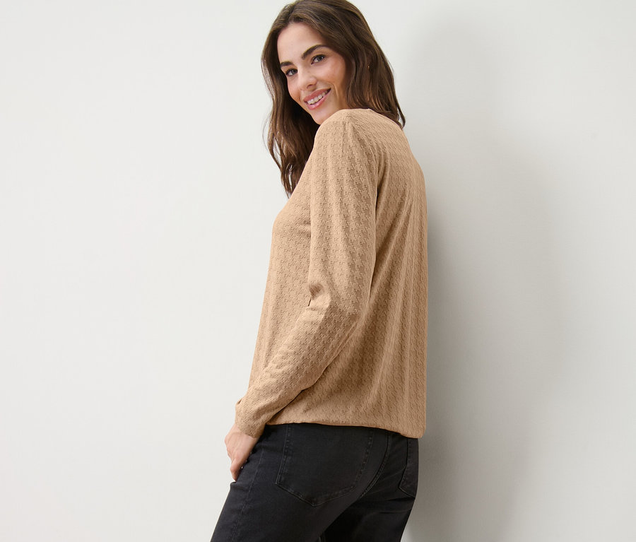 Eine lächelnde Frau blickt über ihre Schulter und posiert in einem strukturierten Langarmshirt in Camel und schwarzen Jeans.