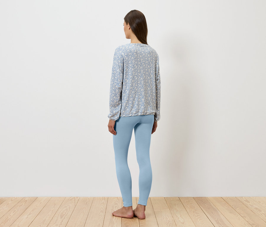 Frau steht mit dem Rücken zum Betrachter, trägt blaue Leggings und eine gemusterte Bluse.