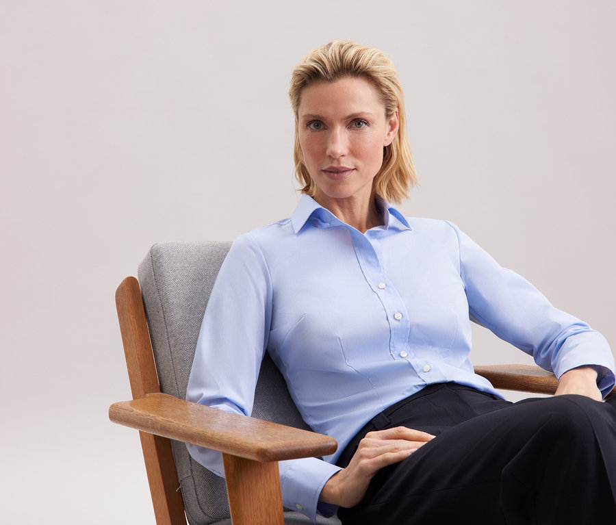 Blonde Frau sitzt in einem grauen Sessel, trägt ein blaues Hemd und eine schwarze Hose.