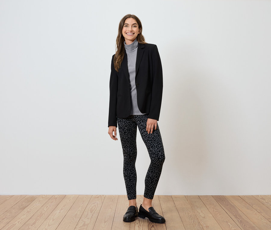 Frau in schwarzem Blazer, grauem Rollkragenpullover und gemusterten Leggings steht auf Holzboden.