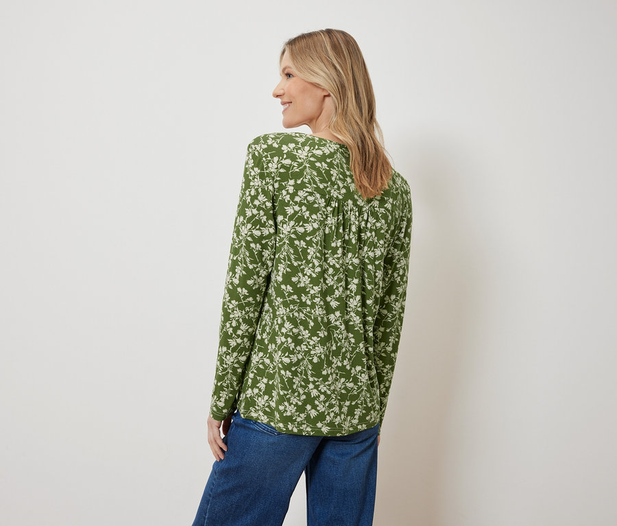 Rückansicht einer blonden Frau in einer grünen Bluse mit Blumenmuster und blauen Jeans.