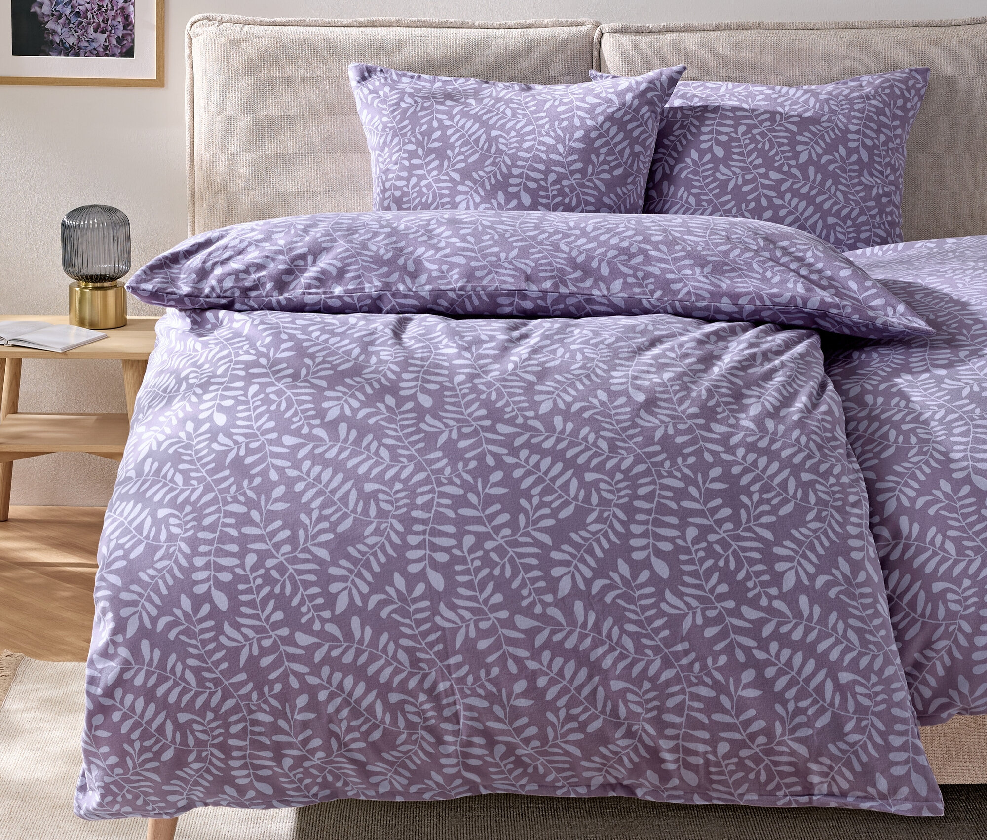 Bettwäsche mit lila Blättermotiv auf einem Bett mit beigem Kopfteil.