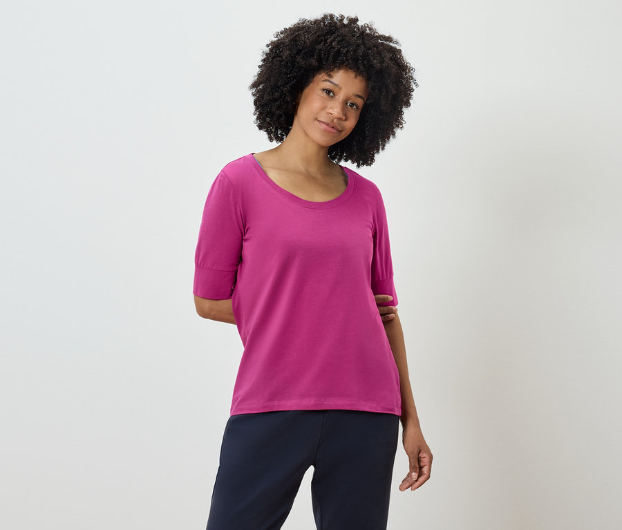 Frau mit Afro-Frisur posiert in einem pinkfarbenen T-Shirt und dunkelblauen Hosen.