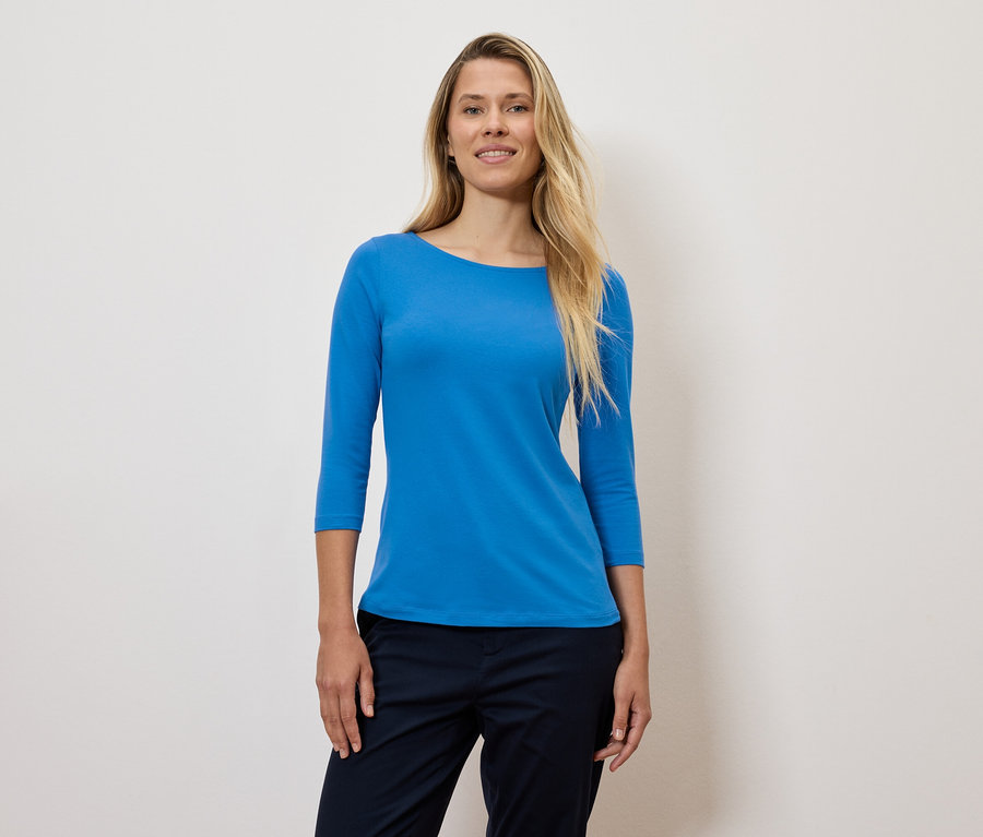 Eine Frau mit blonden Haaren trägt ein blaues Dreiviertelarmshirt und eine dunkelblaue Hose.