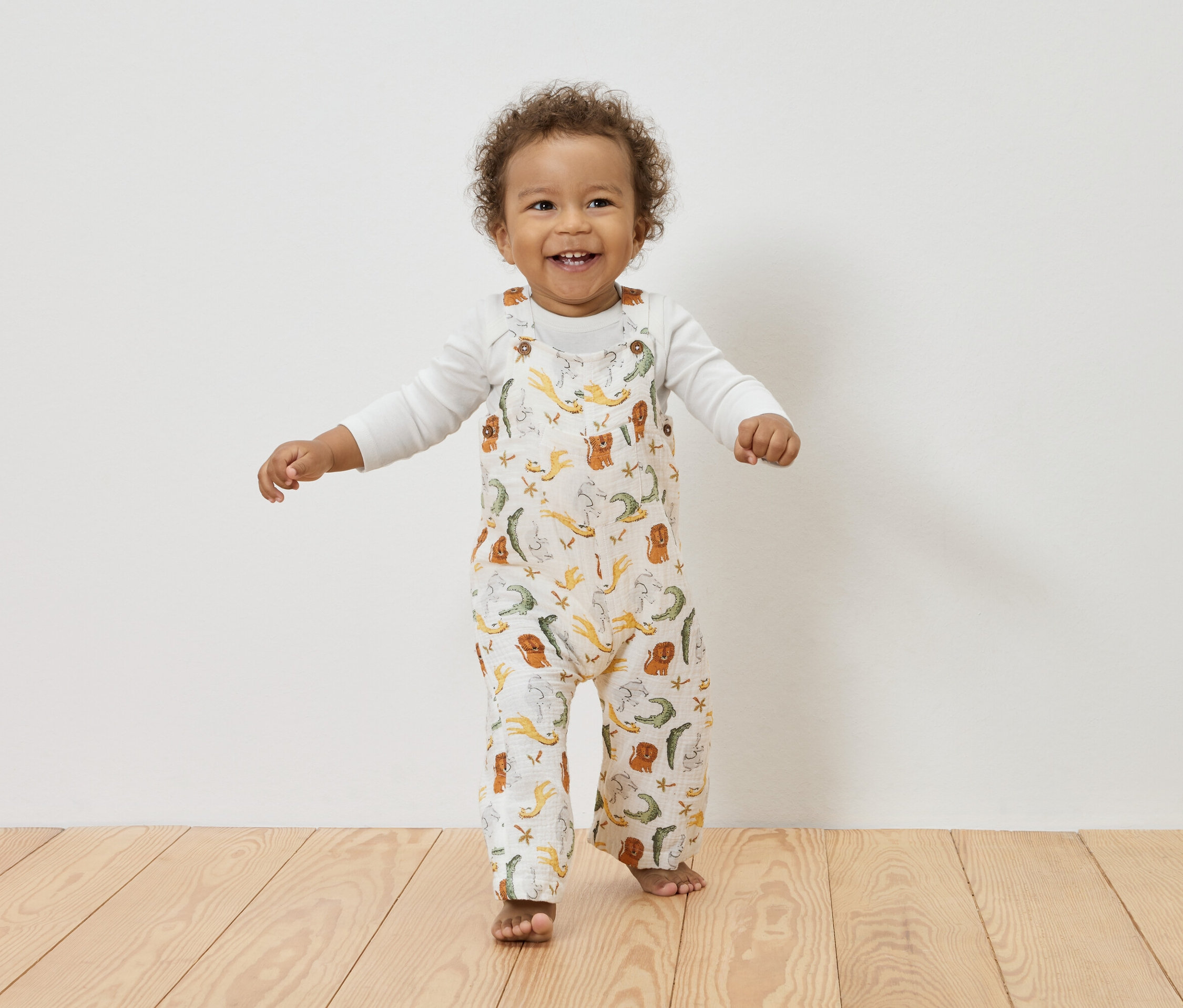 Lächelndes Kleinkind mit lockigem Haar steht auf einem Holzboden und trägt ein weißes Langarmshirt unter einem weißen Overall mit Tiermuster.