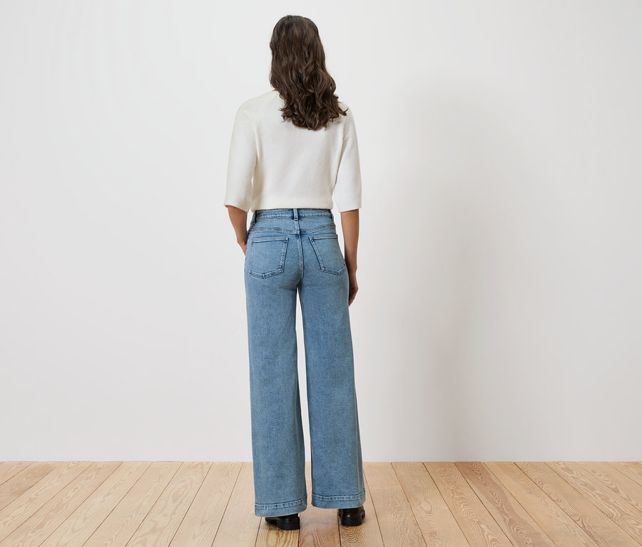 Eine Frau steht mit dem Rücken zur Kamera und trägt eine helle Bluse und weite Jeans.