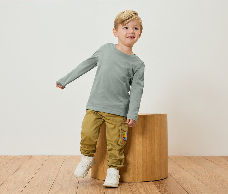 Ein blonder Junge in einem grünen Langarmshirt und Khaki-Hosen steht neben einem Holzhocker.