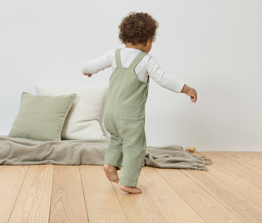 Kleinkind in grüner Latzhose balanciert auf Holzboden neben Kissen und Decke.