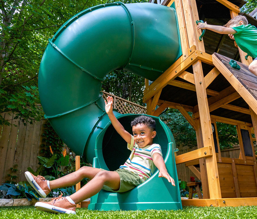 Ein Kind rutscht die grüne Röhrenrutsche des Spielturms »Bolzo« hinunter, während ein anderes Kind auf den Spielturm klettert.