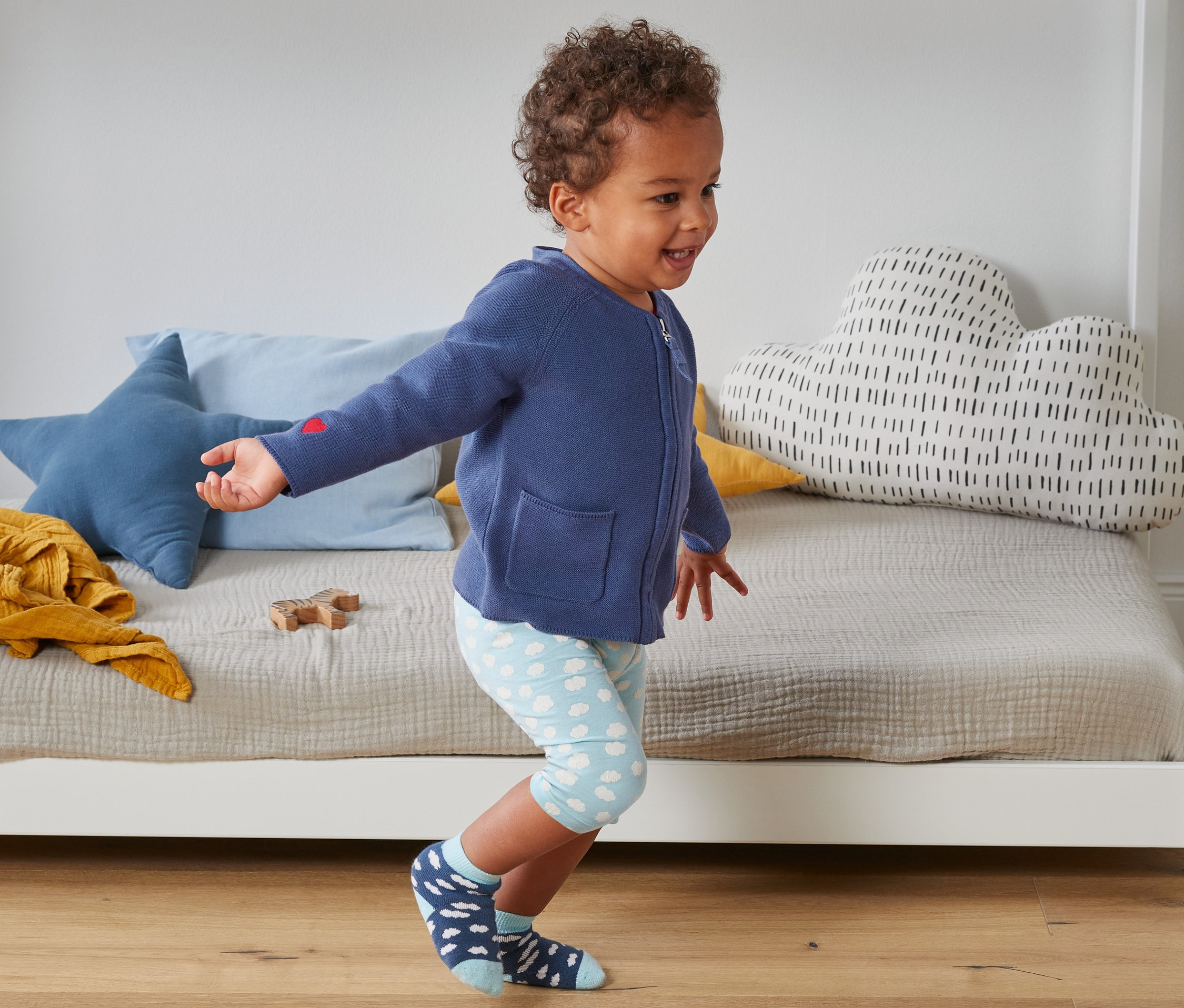 Ein Kind geht in einer blauen Strickjacke aus Bio-Baumwolle und Socken mit Wolken vor einem Kinderbett.