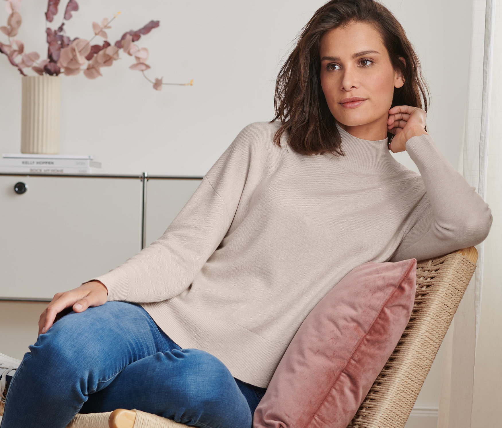Frau sitzt im Sessel und trägt einen Strickpullover mit Stehkragen.
