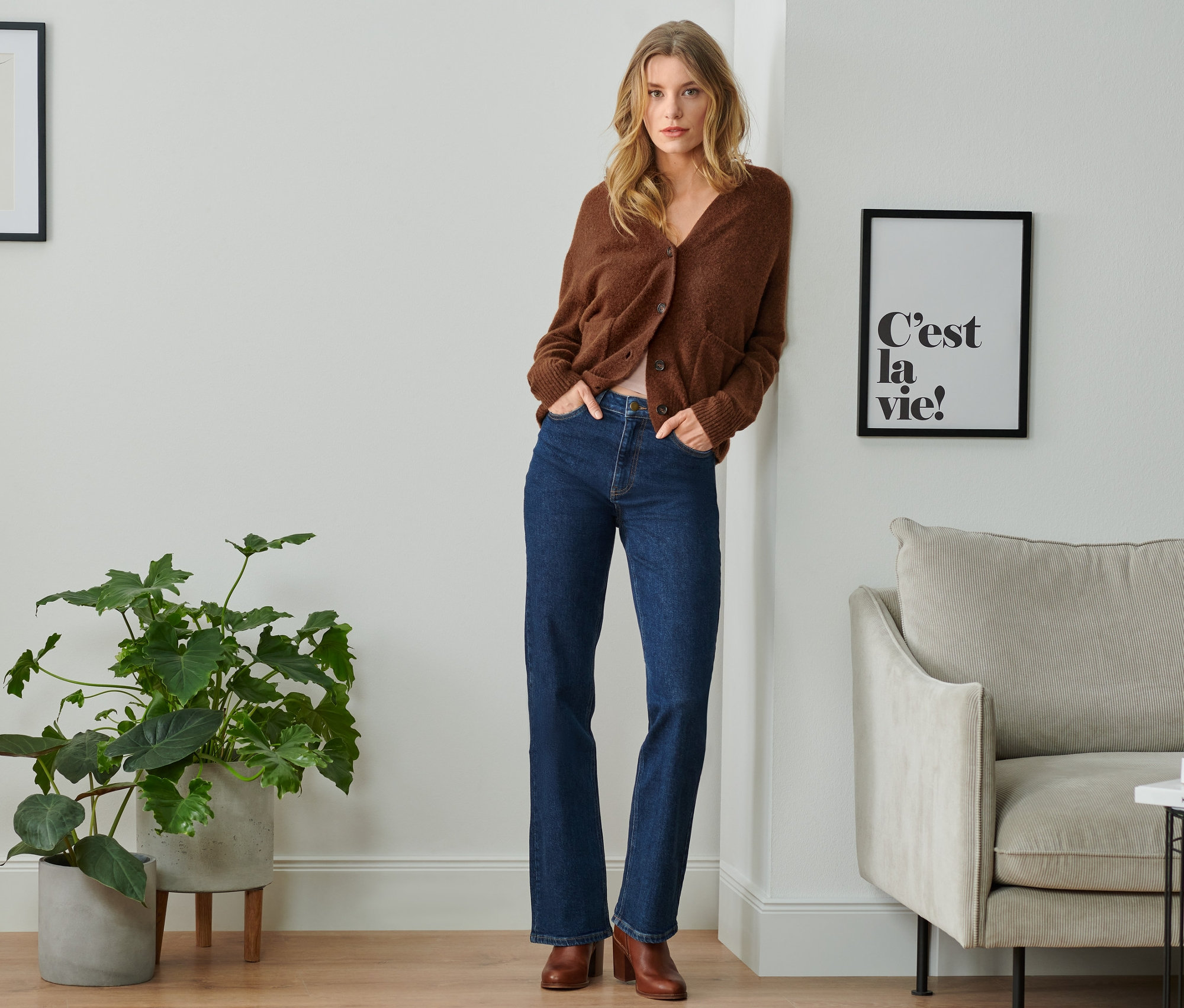 Eine Frau lehnt an einer Wand und trägt eine braune Strickjacke und eine Bootcut-Jeans.