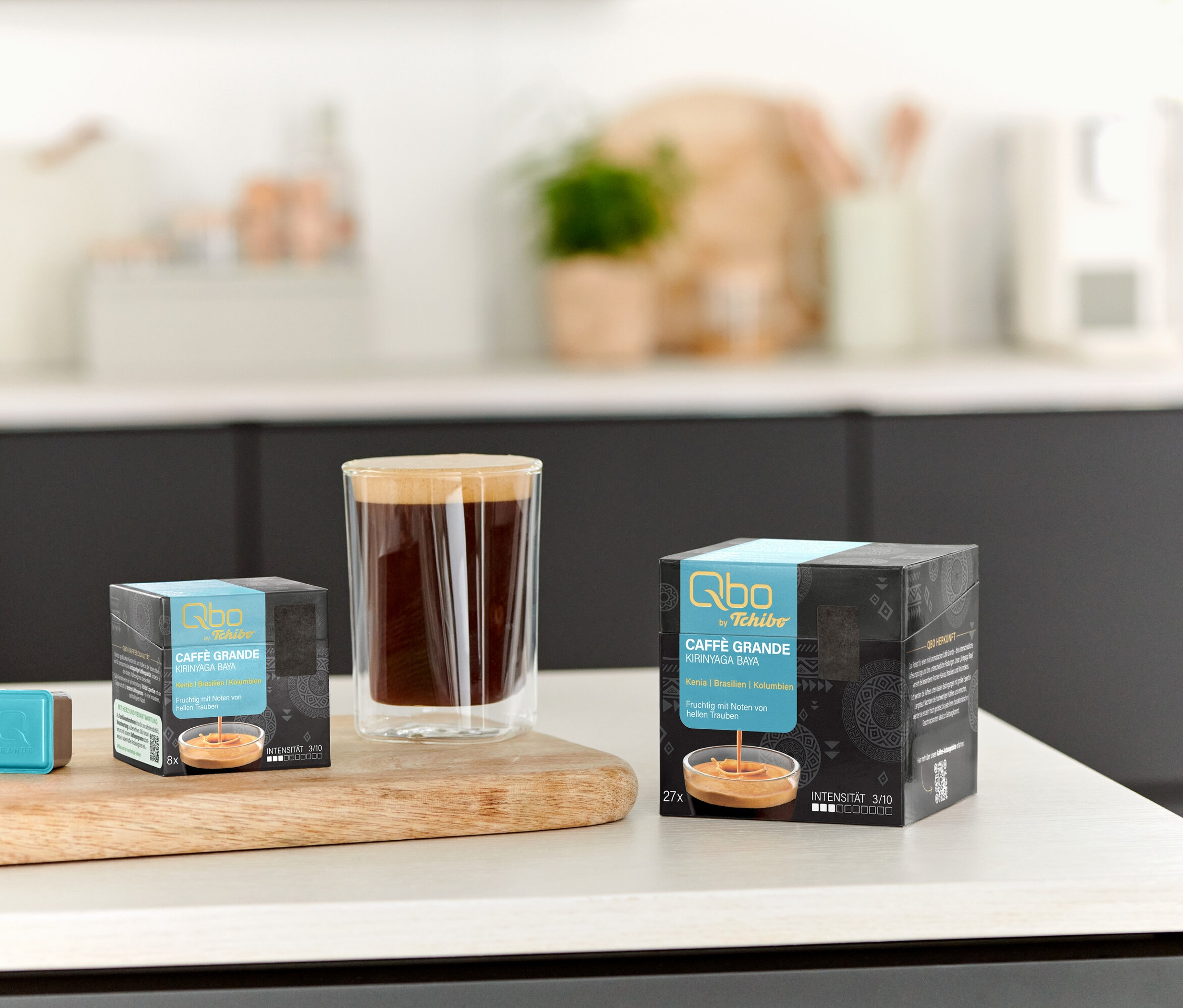 Auf einem Tisch stehen eine Tasse Kaffee, ein Qbo Caffè Grande KIRINYAGA BAYA - 8 Kapseln und ein Qbo Caffè Grande KIRINYAGA BAYA - 27 Kapseln.