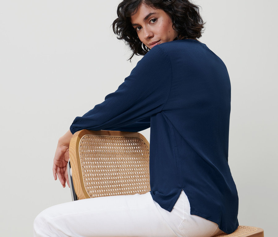 Frau sitzt seitlich auf einem Stuhl und trägt eine blaue Tunika-Bluse.