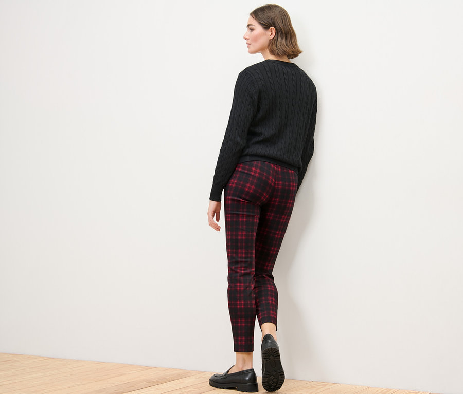 Frau steht mit dem Rücken zur Kamera und trägt eine karierte Stretchhose, einen schwarzen Pullover und schwarze Schuhe.