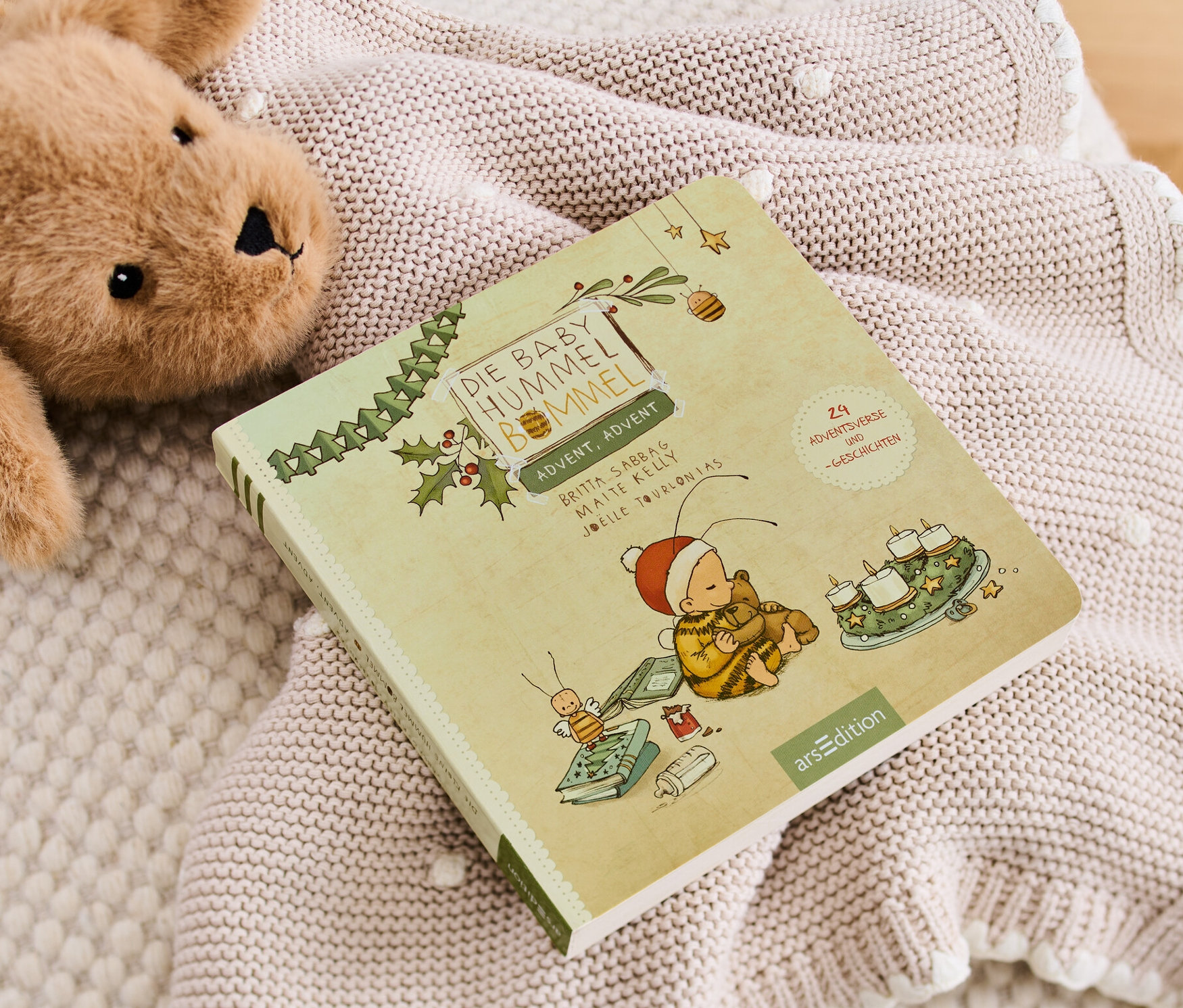 Teddybär und Buch »Die Baby-Hummel Bommel ‒ Advent, Advent« liegen auf einer Strickdecke.