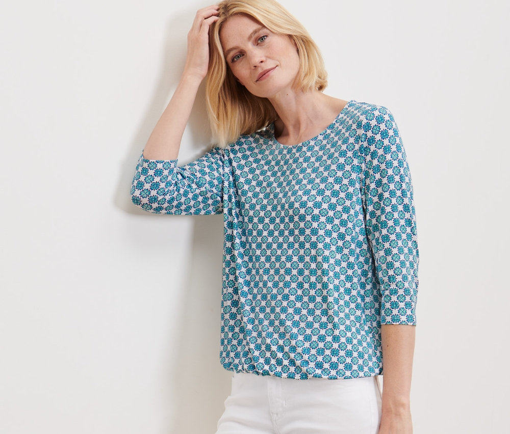 Frau posiert an einer Wand in einem Blusenshirt mit Alloverprint und einer weißen Hose.