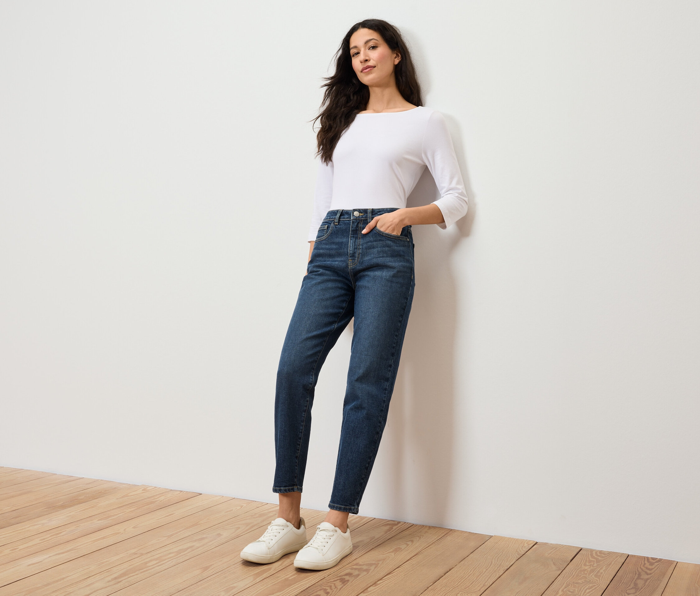 Eine Frau in einer weißen Bluse und blauen Jeans steht an einer weißen Wand.