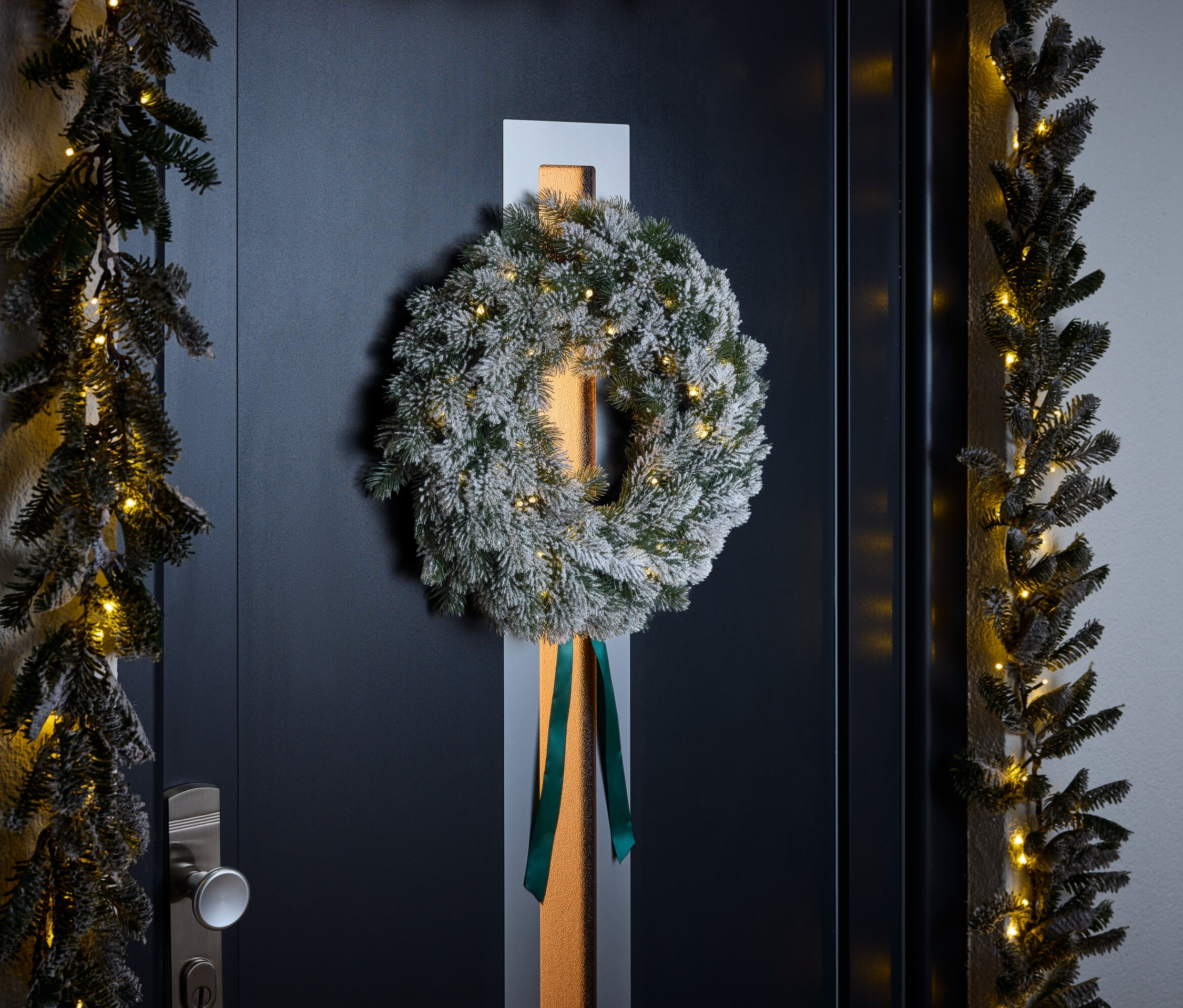 Ein mit weißem Pulver bedeckter Kranz mit grünem Band hängt an einer dunkelblauen Tür. Girlanden mit kleinen Lichtern säumen die Tür.